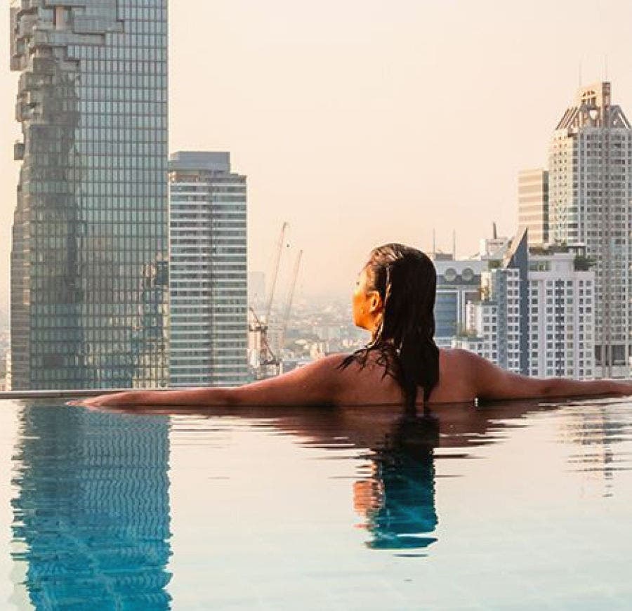 A brunette lady stands facing away, inside a swimming pool, on top of a luxury building, in the center of a modern city.